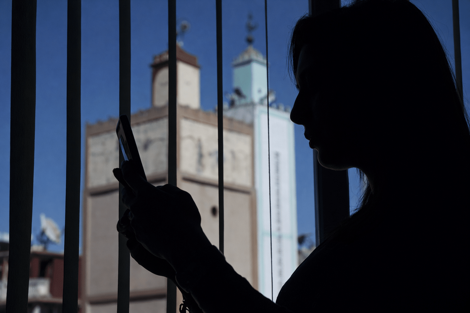 Jeune femme marocaine utilisant son smartphone devant une fenêtre, illustrant l’impact des réseaux sociaux sur le bien-être